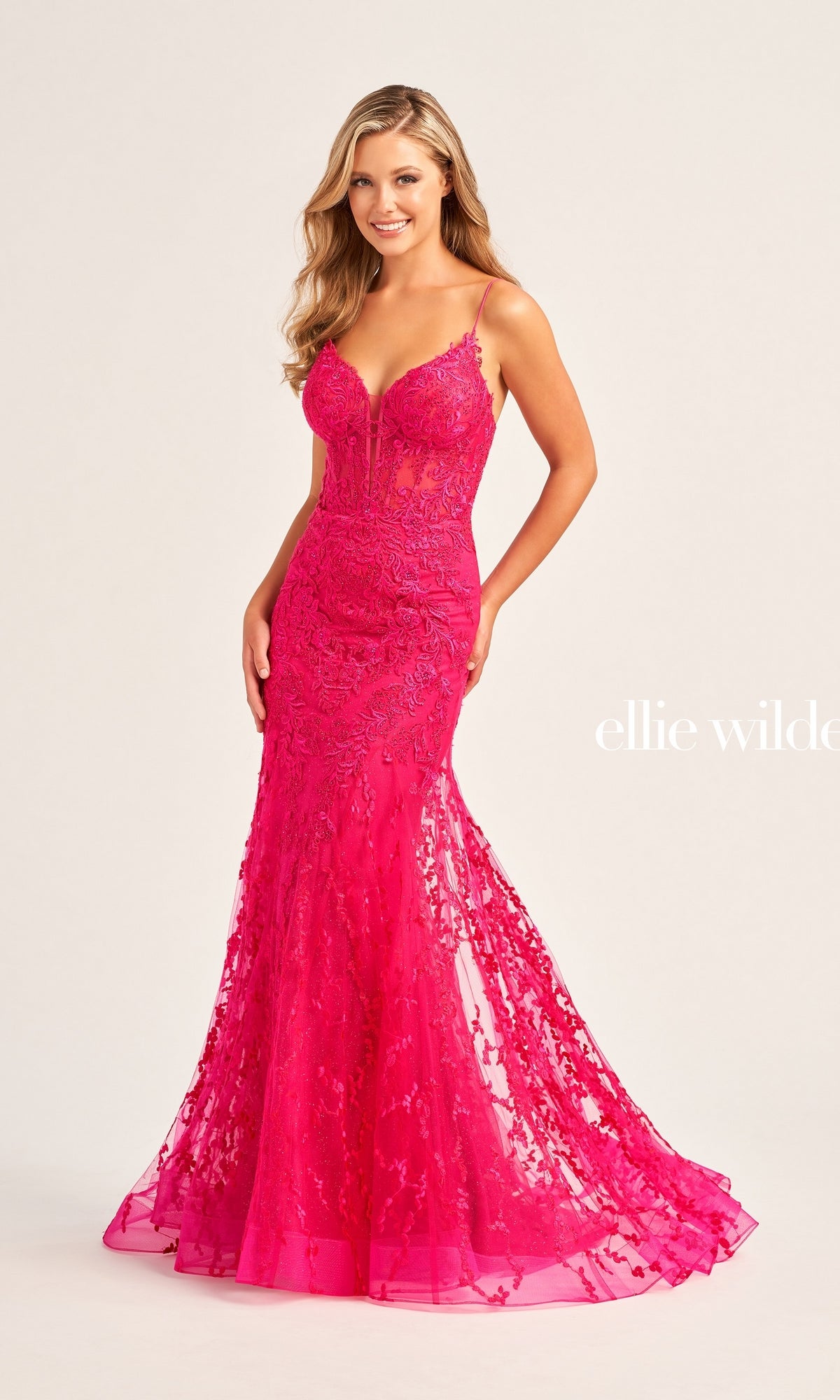 A woman wearing the Ellie Wilde Beaded-Tulle Long Prom Dress EW35010 in bright pink, featuring lace embellishments, thin straps, and delicate beaded embroidery, poses against a plain light background.